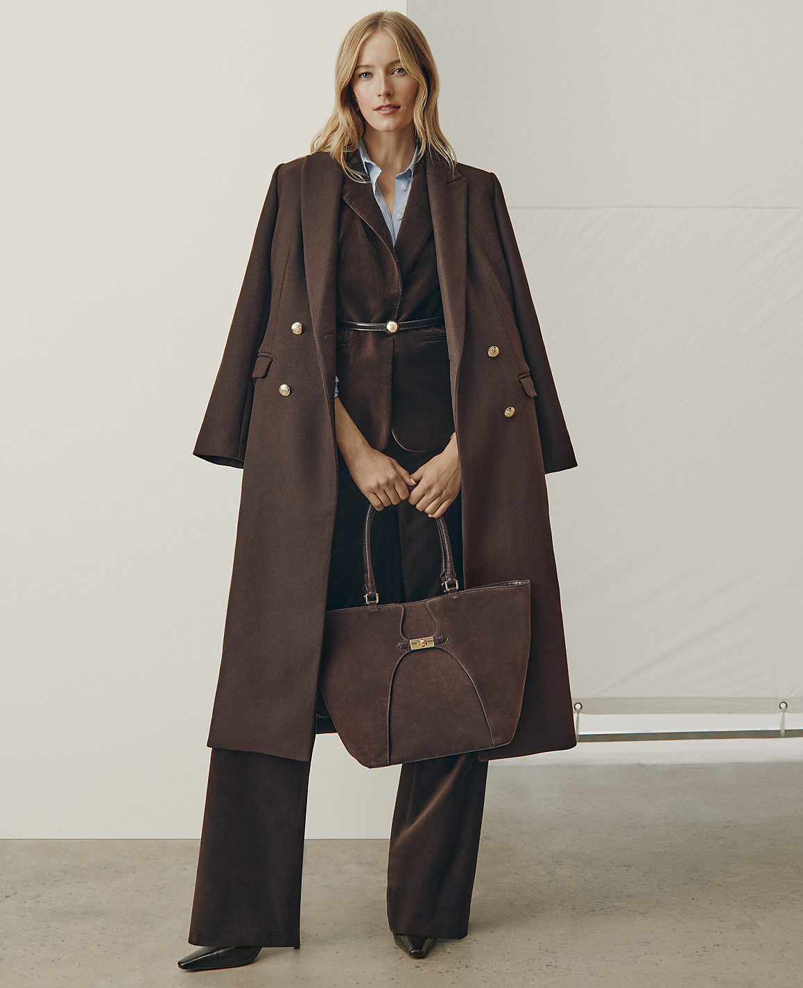A woman wearing a brown coat and matching suit stands holding a brown handbag against a plain white background.