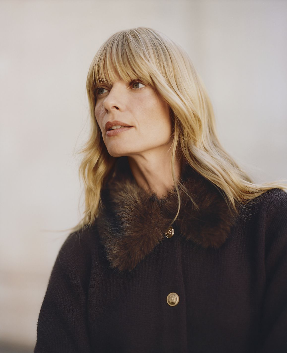 A blonde woman wearing a dark coat with a faux-fur collar stands against a blurred, neutral-toned background.