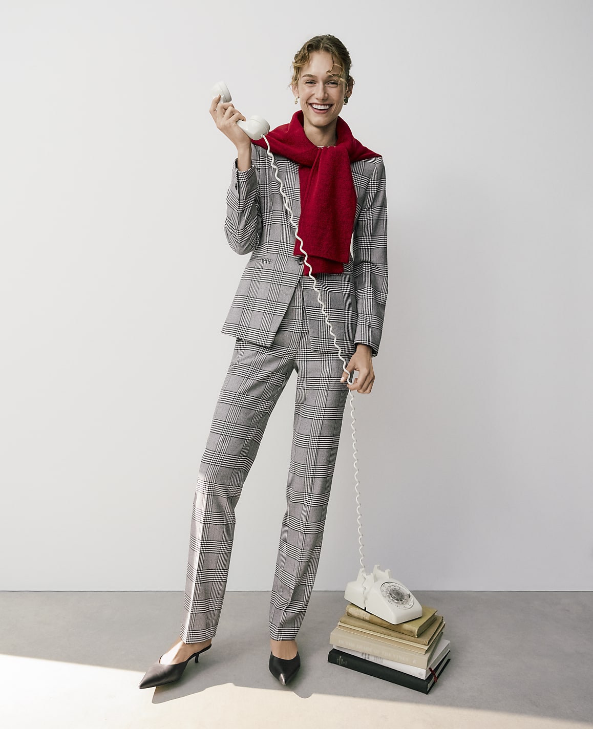 A smiling woman in a plaid suit and red scarf talks on a vintage white telephone resting on a stack of books against a plain white background.