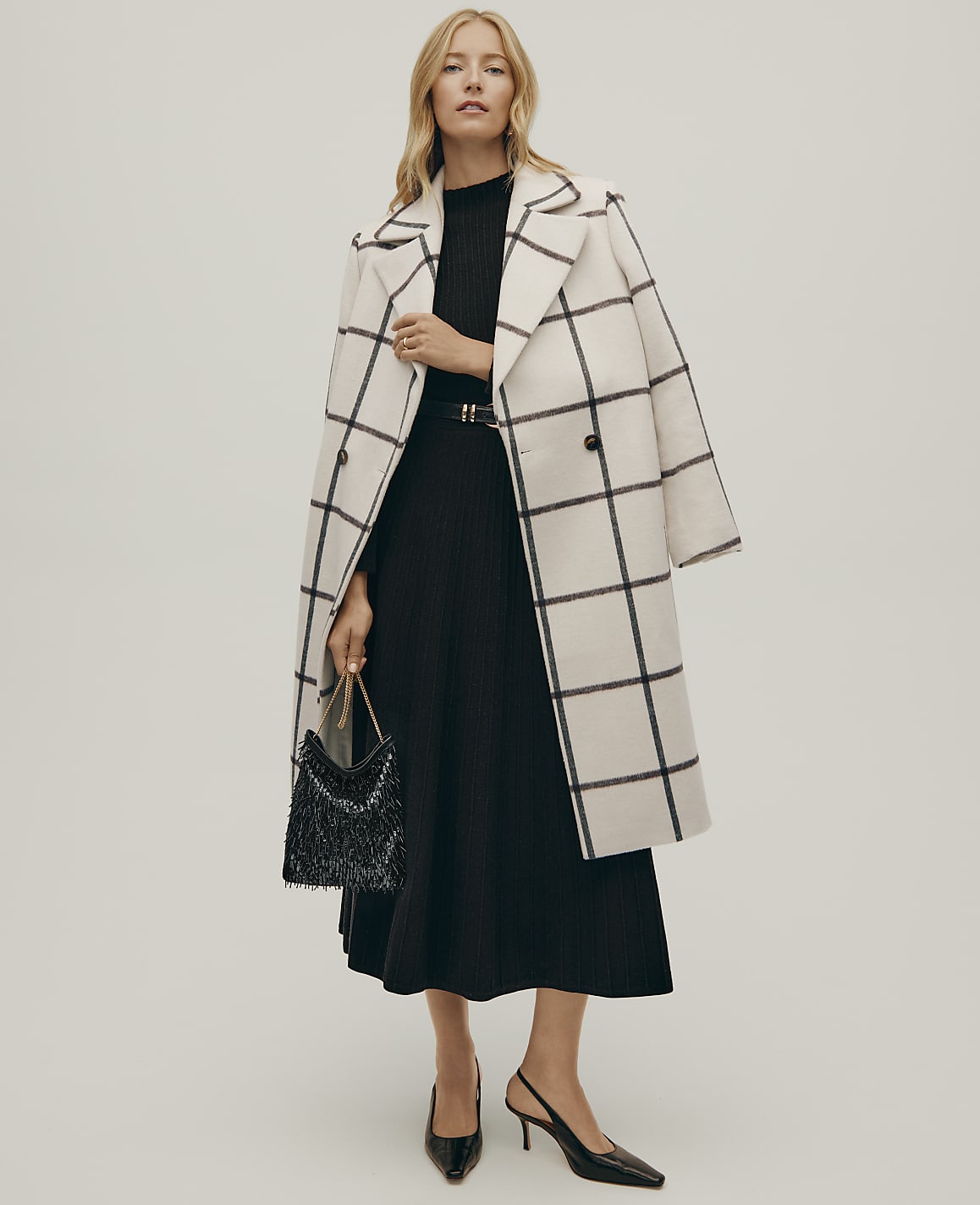 A blonde woman wearing a black pleated dress and a white grid-patterned coat stands against a plain light gray background.