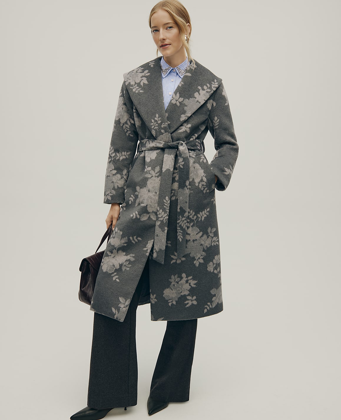 A woman wearing a grey floral-patterned wrap coat and wide-leg trousers stands against a plain, light grey background.