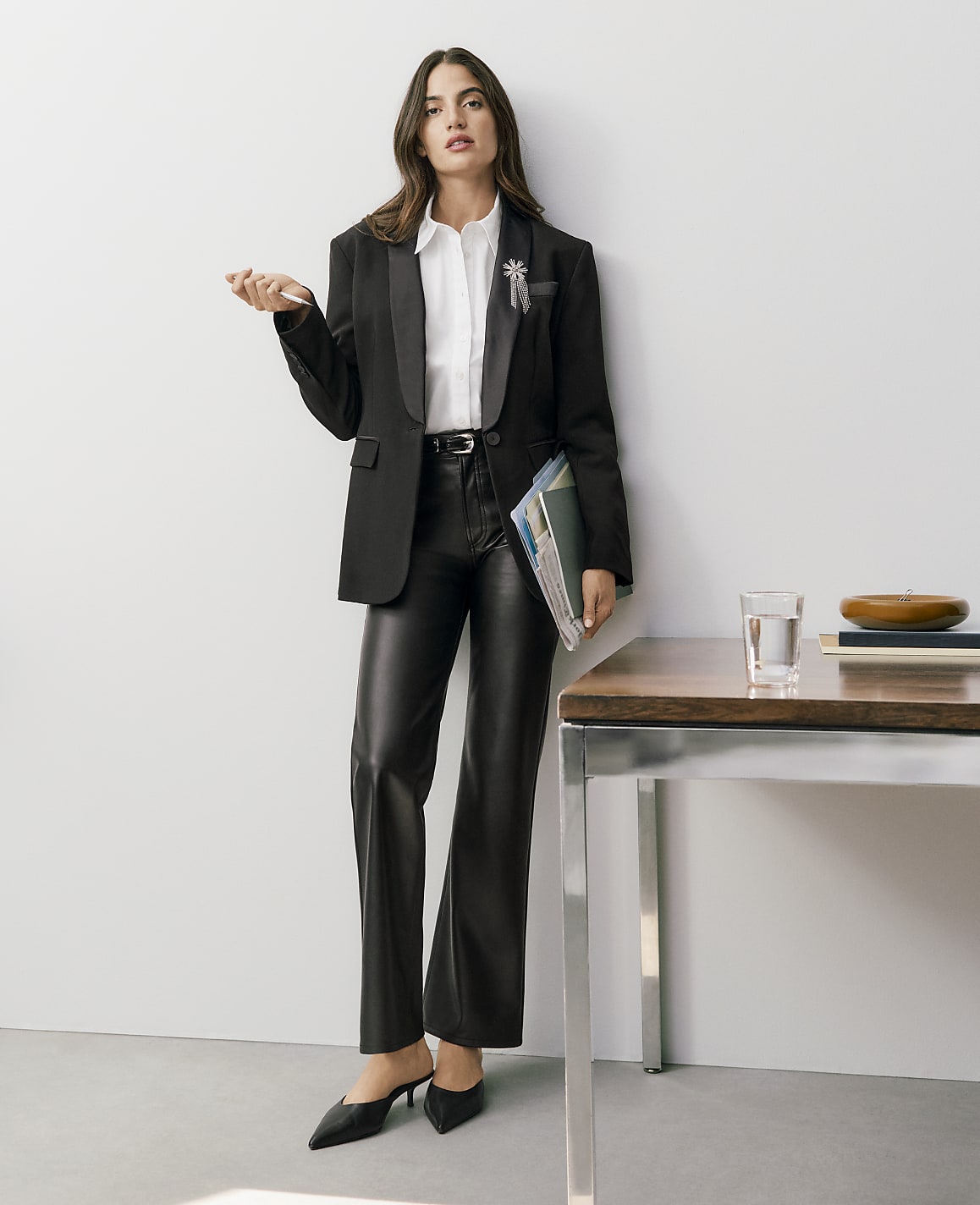 A woman wearing a black blazer, white shirt, and leather trousers stands beside a wooden table against a plain white wall.