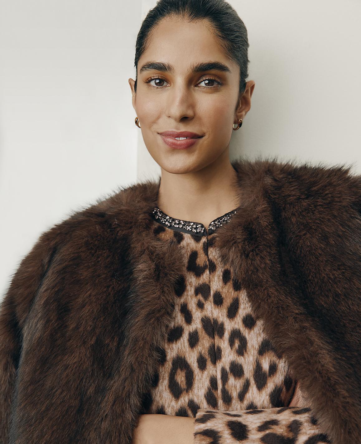 A woman wearing a leopard-print dress and a brown faux-fur coat poses against a plain white background.