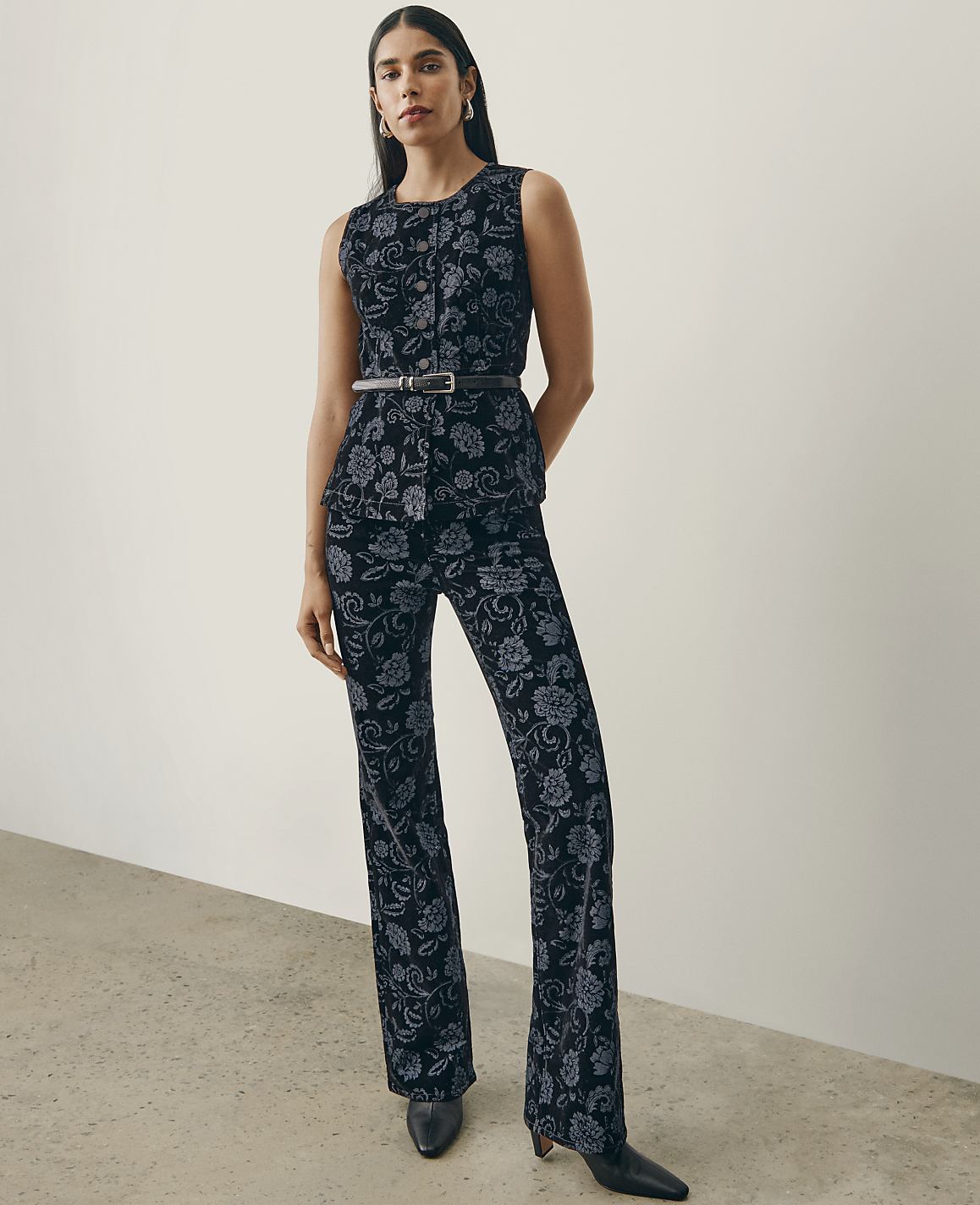 A woman wearing a dark floral print sleeveless top and matching trousers stands against a plain white wall and a light grey floor.