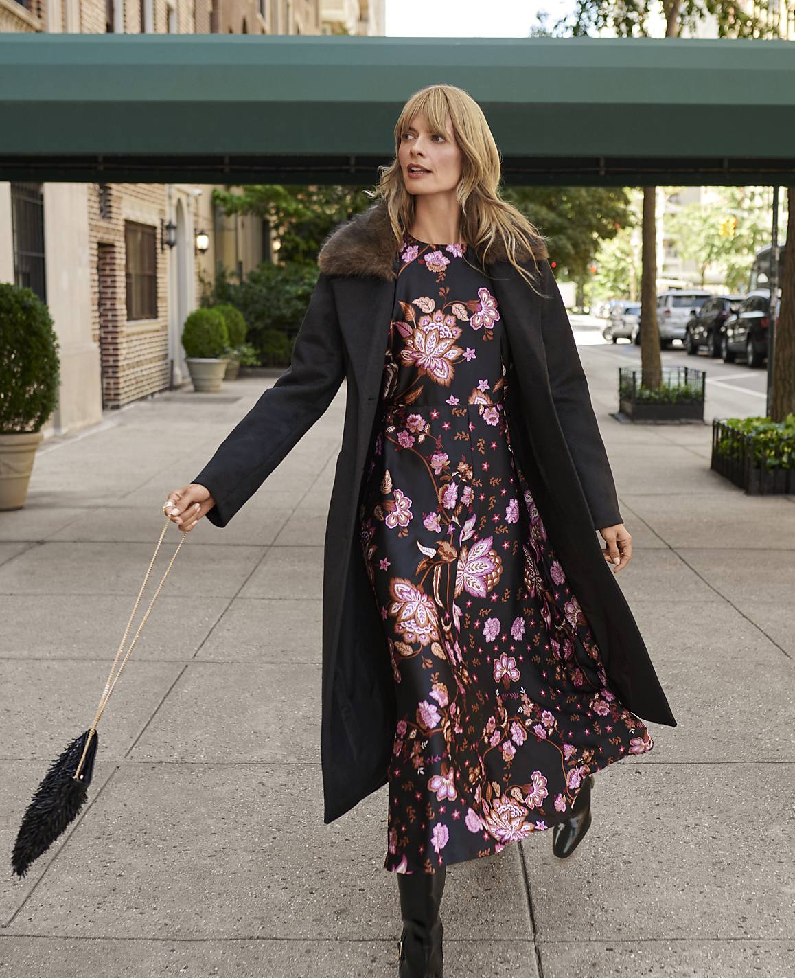 A woman in a floral dress and black coat stands on a city sidewalk with buildings and parked cars in the background.