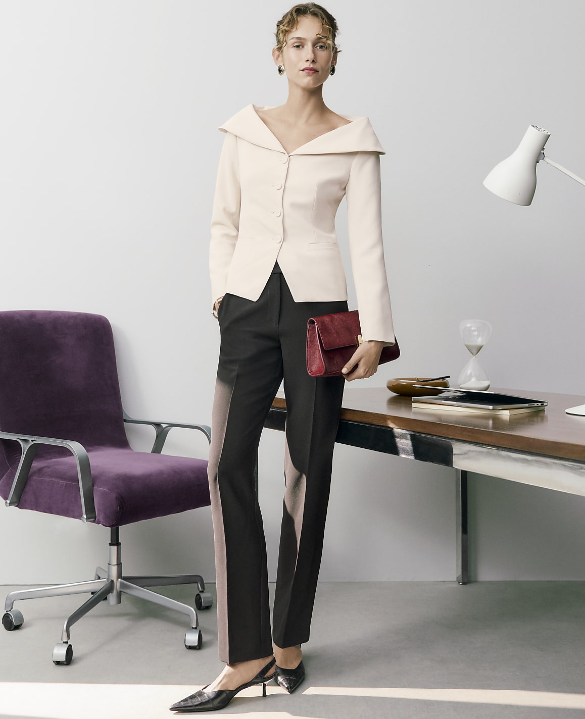 A woman in a pale pink blazer and black trousers stands holding a red clutch in an office setting with a purple chair and a desk.