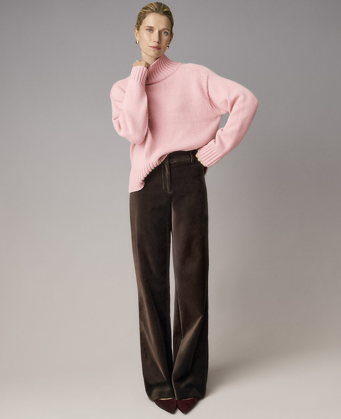 A woman wearing a pink turtleneck sweater and wide-leg brown trousers stands against a plain grey background.