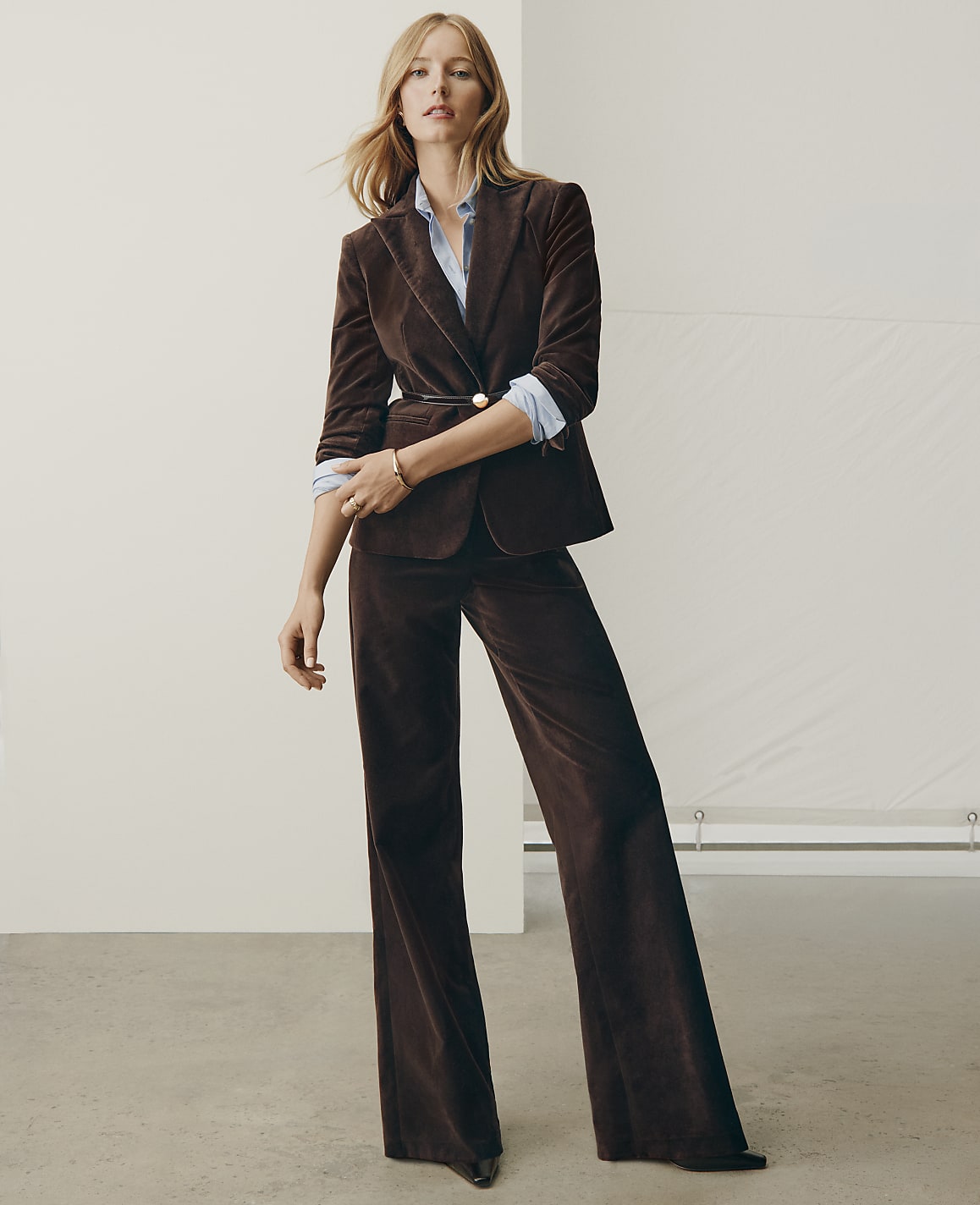 A woman wearing a brown velvet suit and a light blue shirt stands against a plain white studio background.