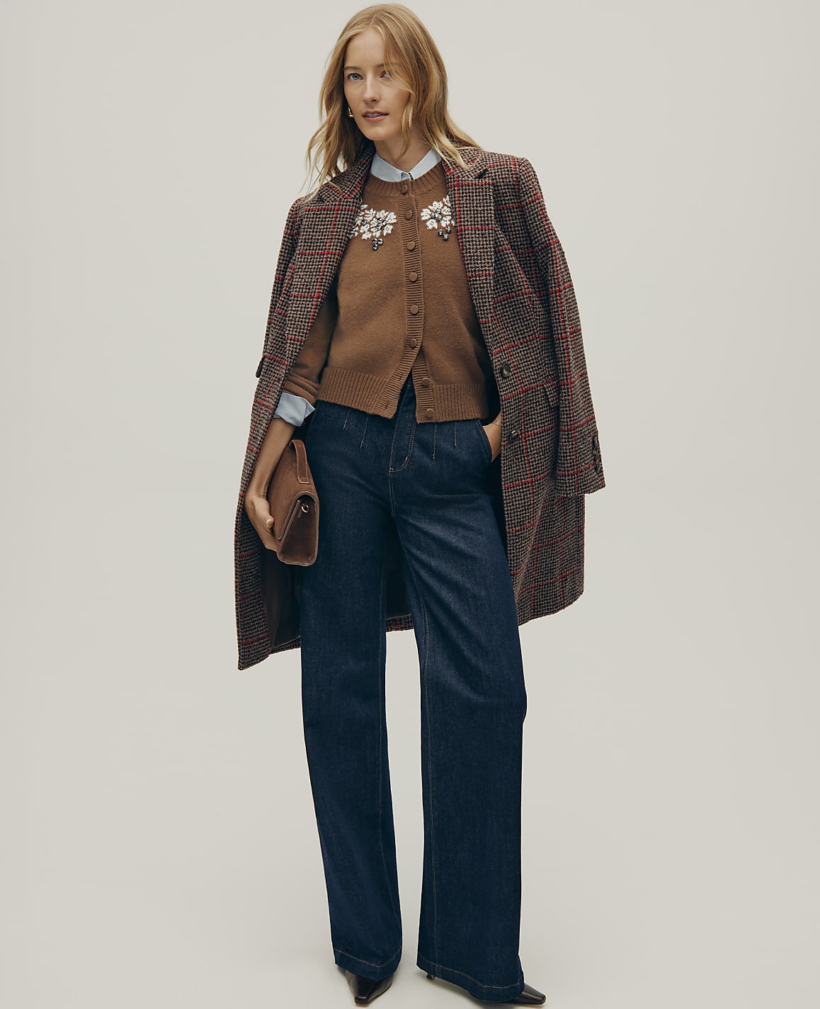 A woman wearing a brown cardigan, wide-leg jeans, and a plaid coat stands against a plain light gray background.