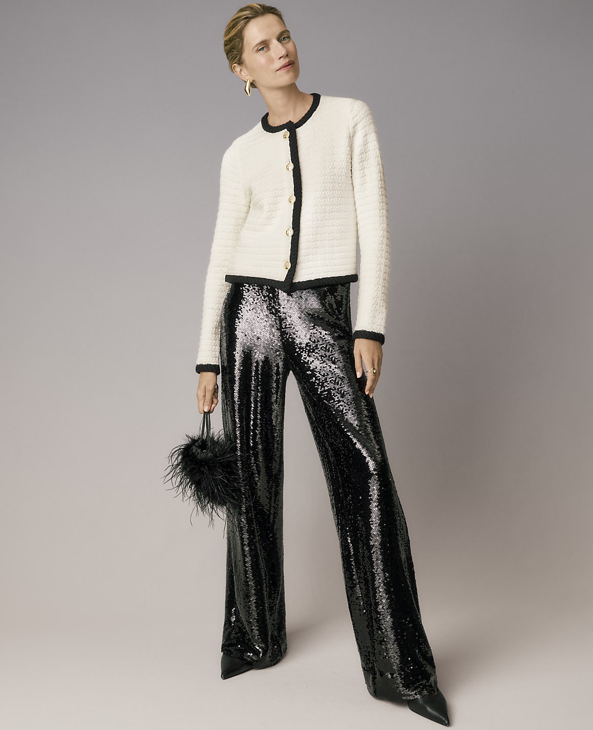 A woman wearing a white tweed jacket and shimmering black sequined trousers stands against a plain grey background.