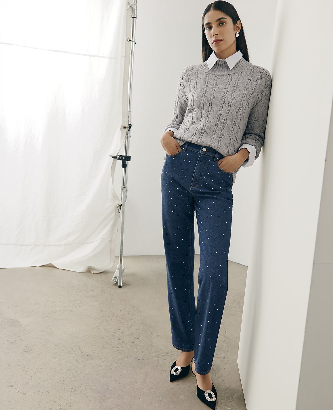 A woman wearing a grey cable-knit sweater and rhinestone-embellished blue jeans leans against a white wall in a studio with a white backdrop.