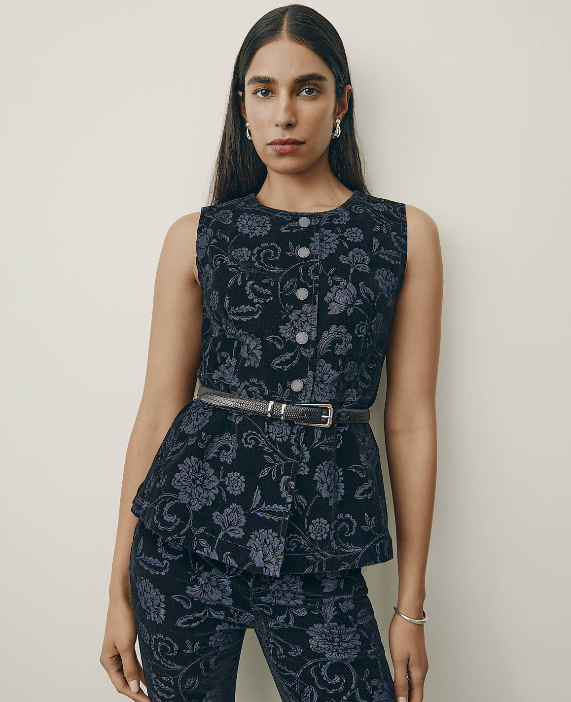 A woman wearing a dark blue floral patterned sleeveless top and matching pants stands against a plain, light-colored wall.