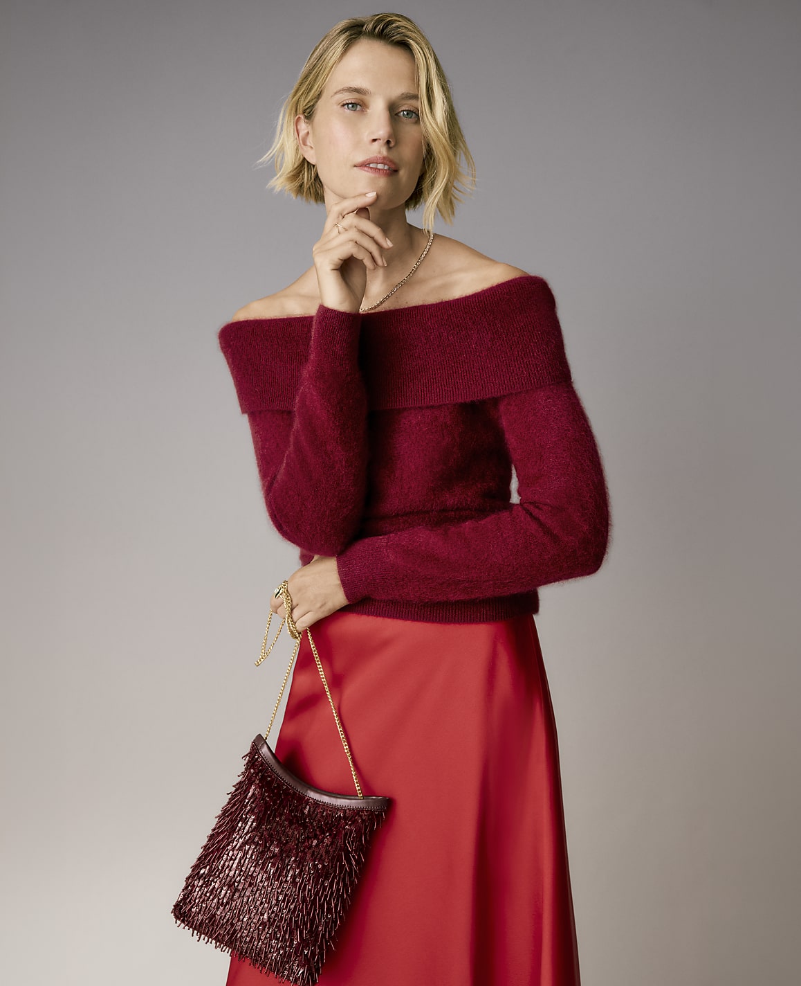 A blonde woman wearing a burgundy off-the-shoulder sweater and a red satin skirt stands against a plain grey background.