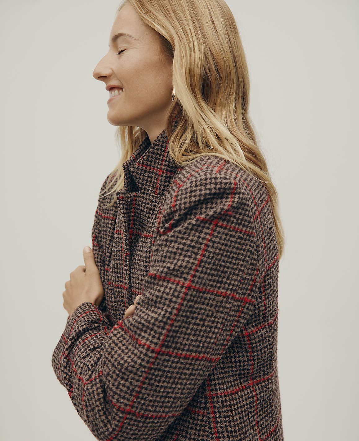 A smiling blonde woman in a brown and red plaid coat stands against a plain, light-colored background.