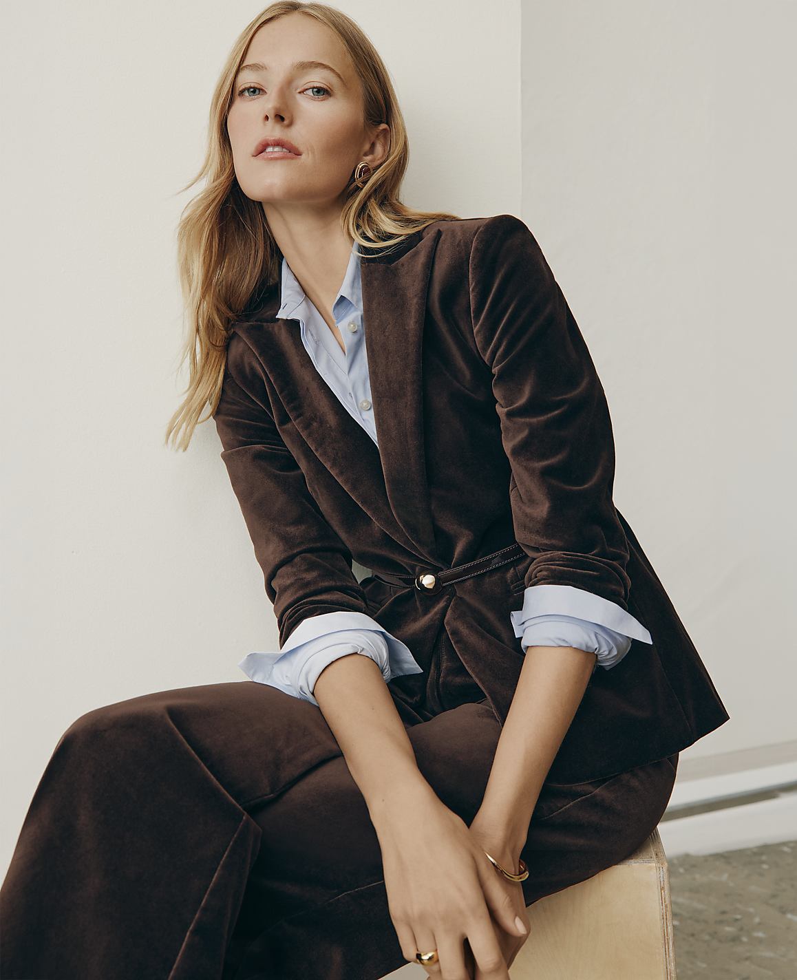 A blonde woman wearing a brown velvet suit and light blue shirt sits against a plain white wall.