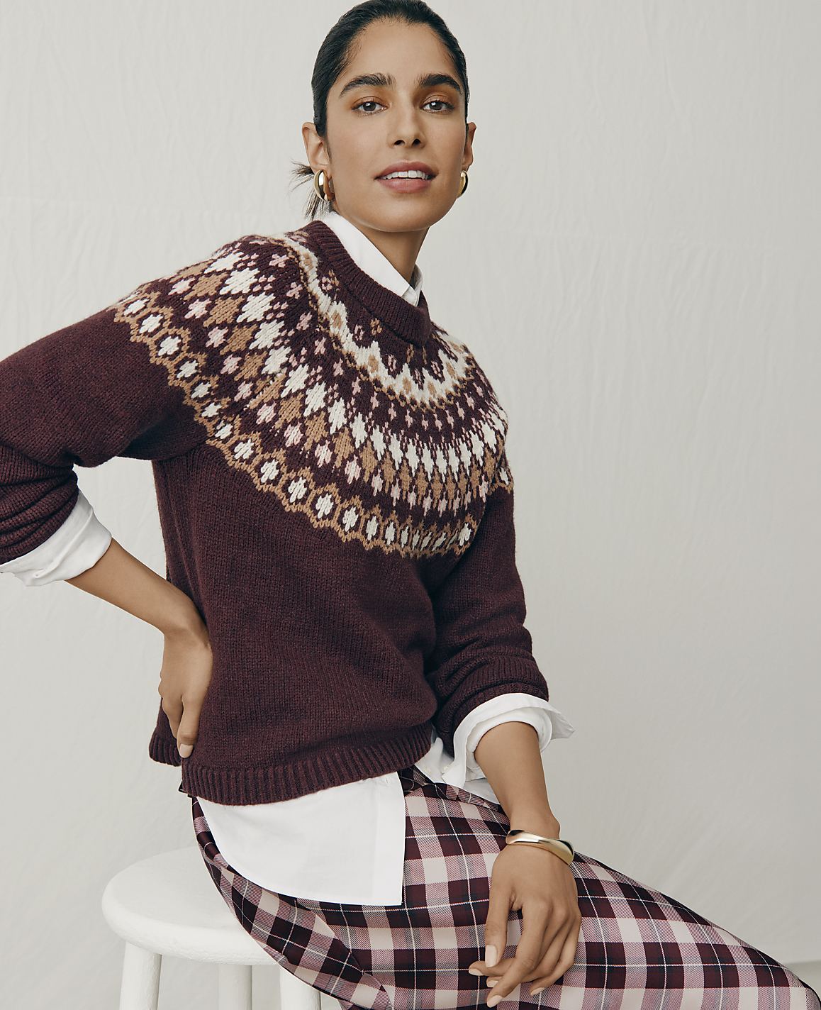 A woman wearing a maroon patterned sweater and plaid pants sits on a white stool against a plain light gray background.