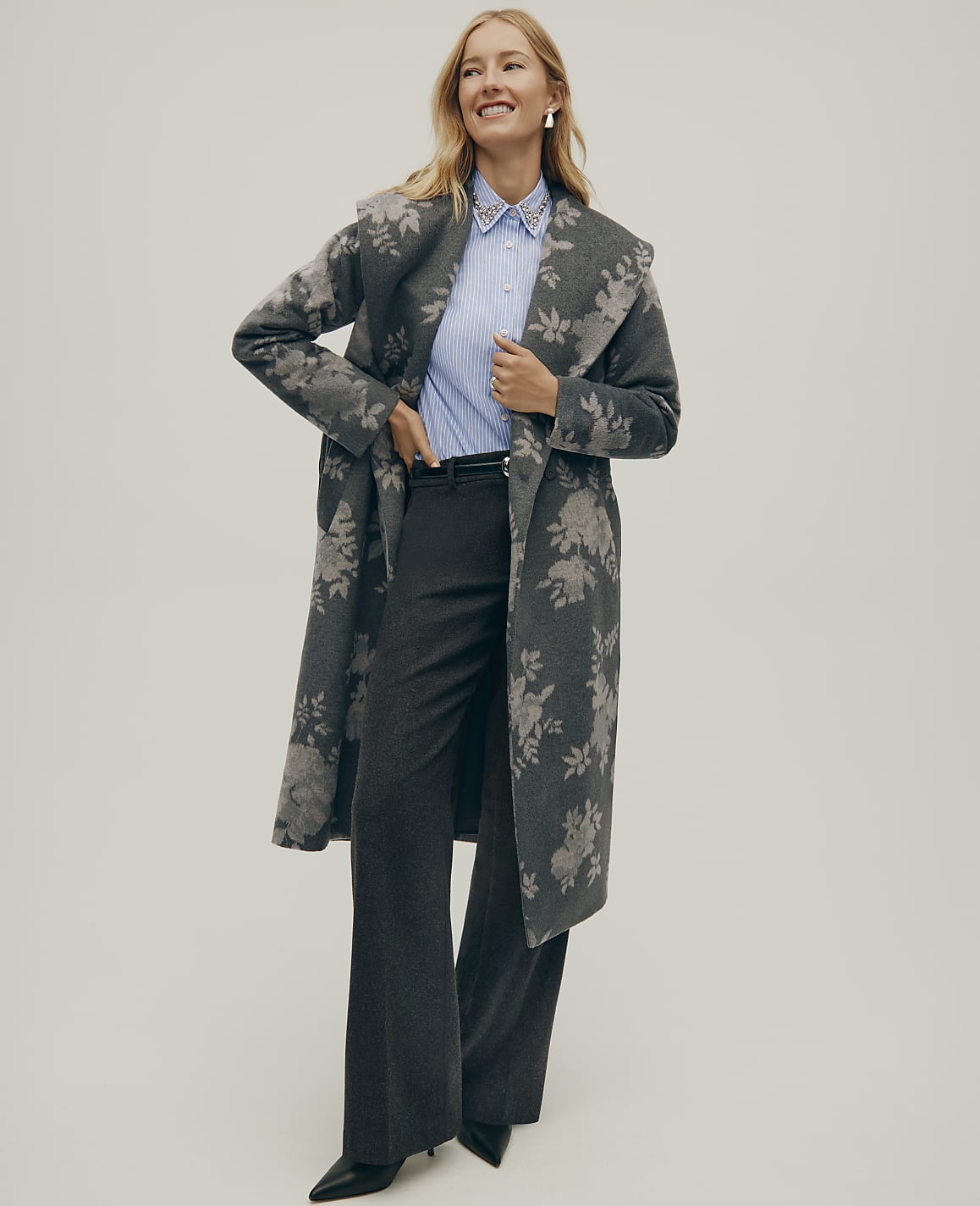 A smiling woman wearing a floral grey coat, a blue striped shirt, and wide-leg trousers stands against a plain light grey background.