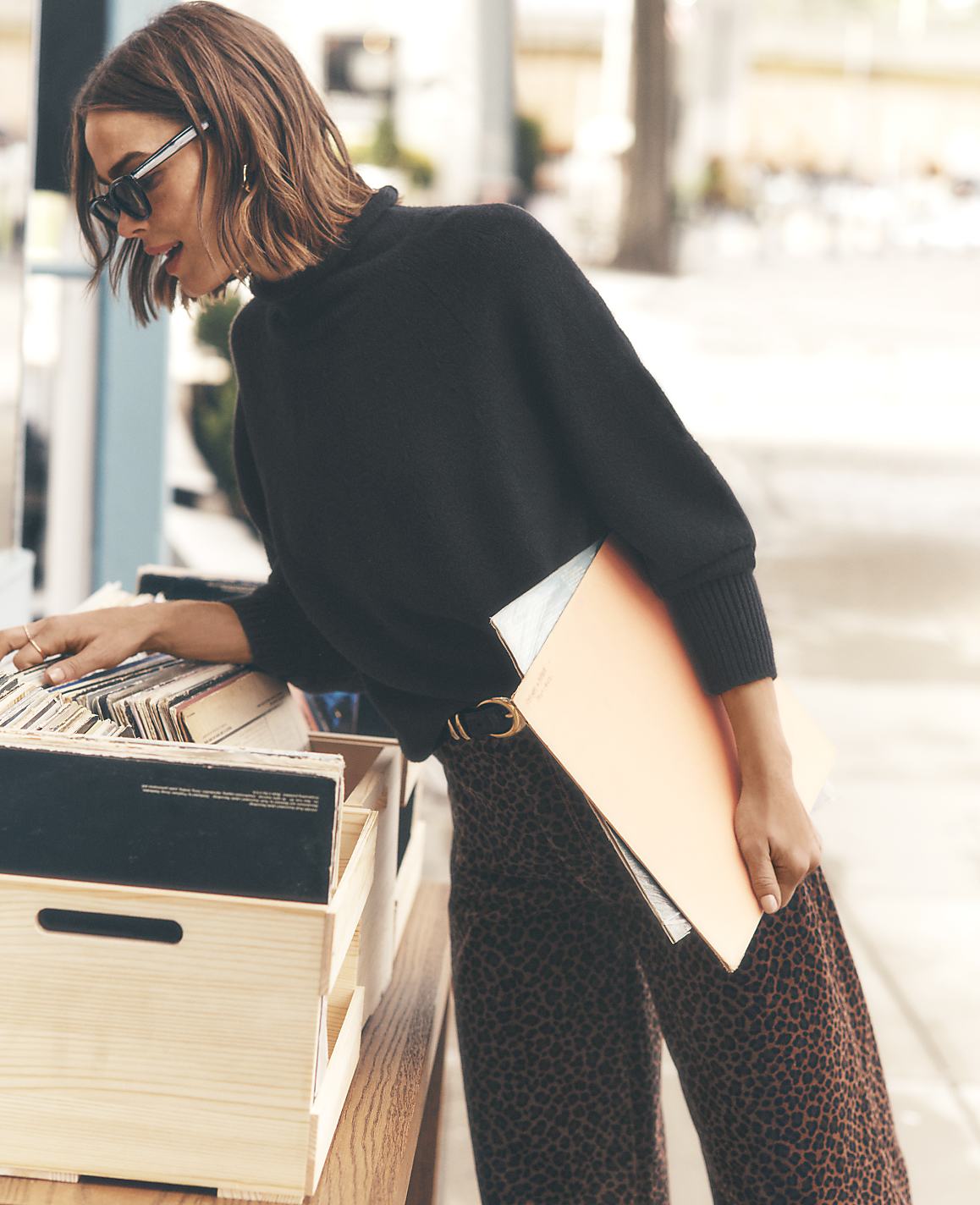 A woman in a black sweater and leopard-print pants browses vinyl records in a wooden crate against a blurred city street background.