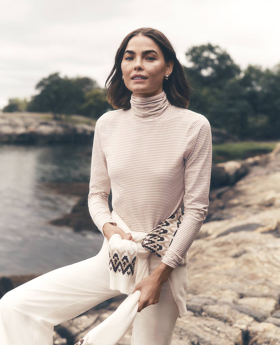 A woman wearing a light-colored turtleneck and white pants stands on a rocky shoreline with a calm body of water and green trees in the background.