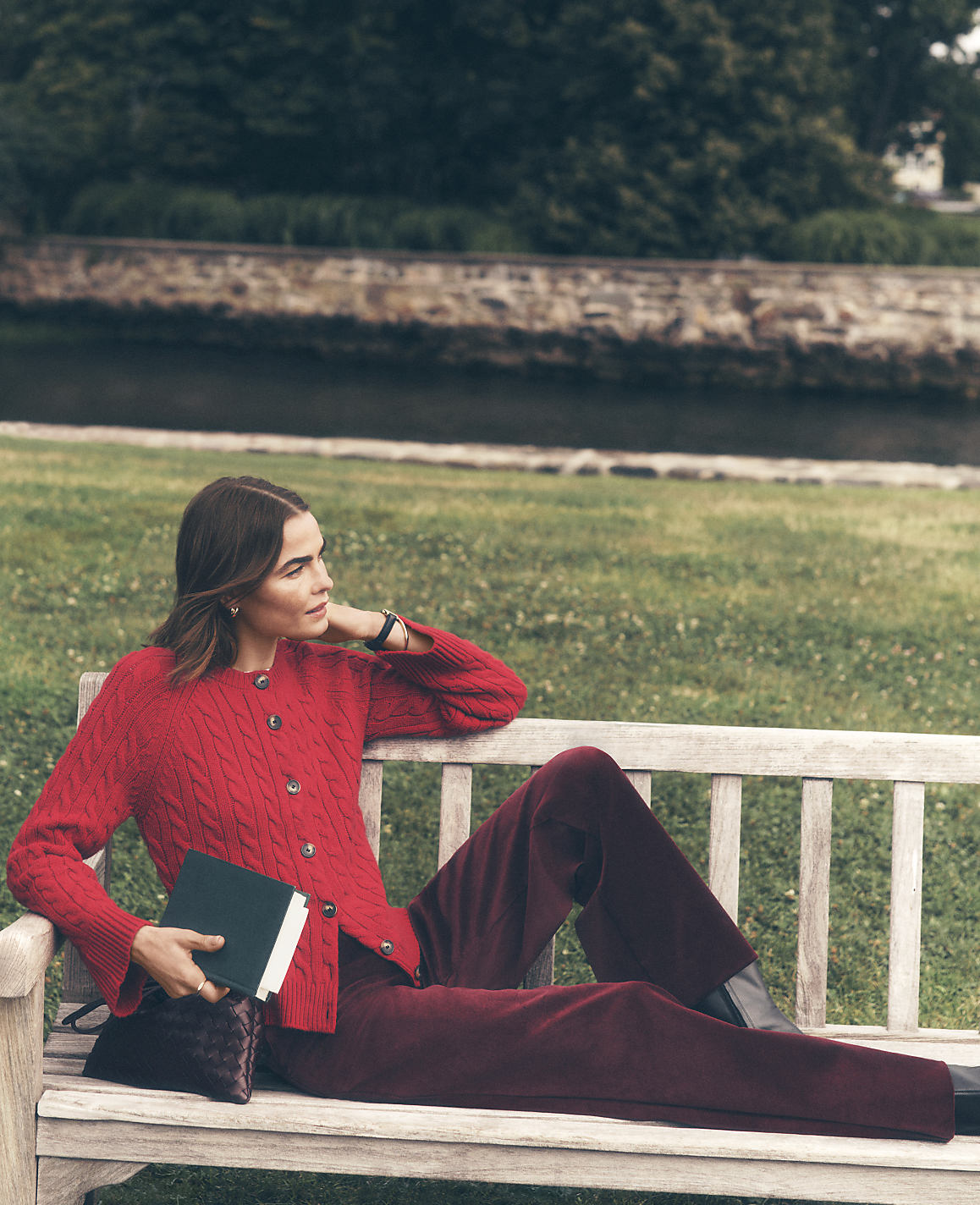 A woman in a red cardigan and burgundy trousers sits on a wooden bench holding a book, with a grassy lawn and a stone riverbank in the background.