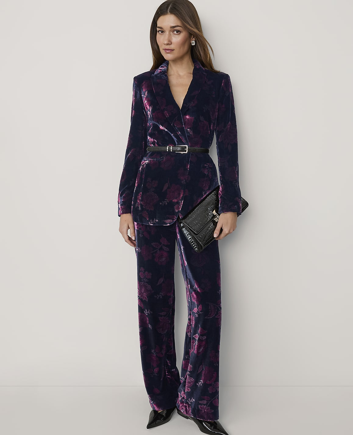 A woman wearing a dark purple velvet floral suit and holding a black clutch stands against a plain white background.