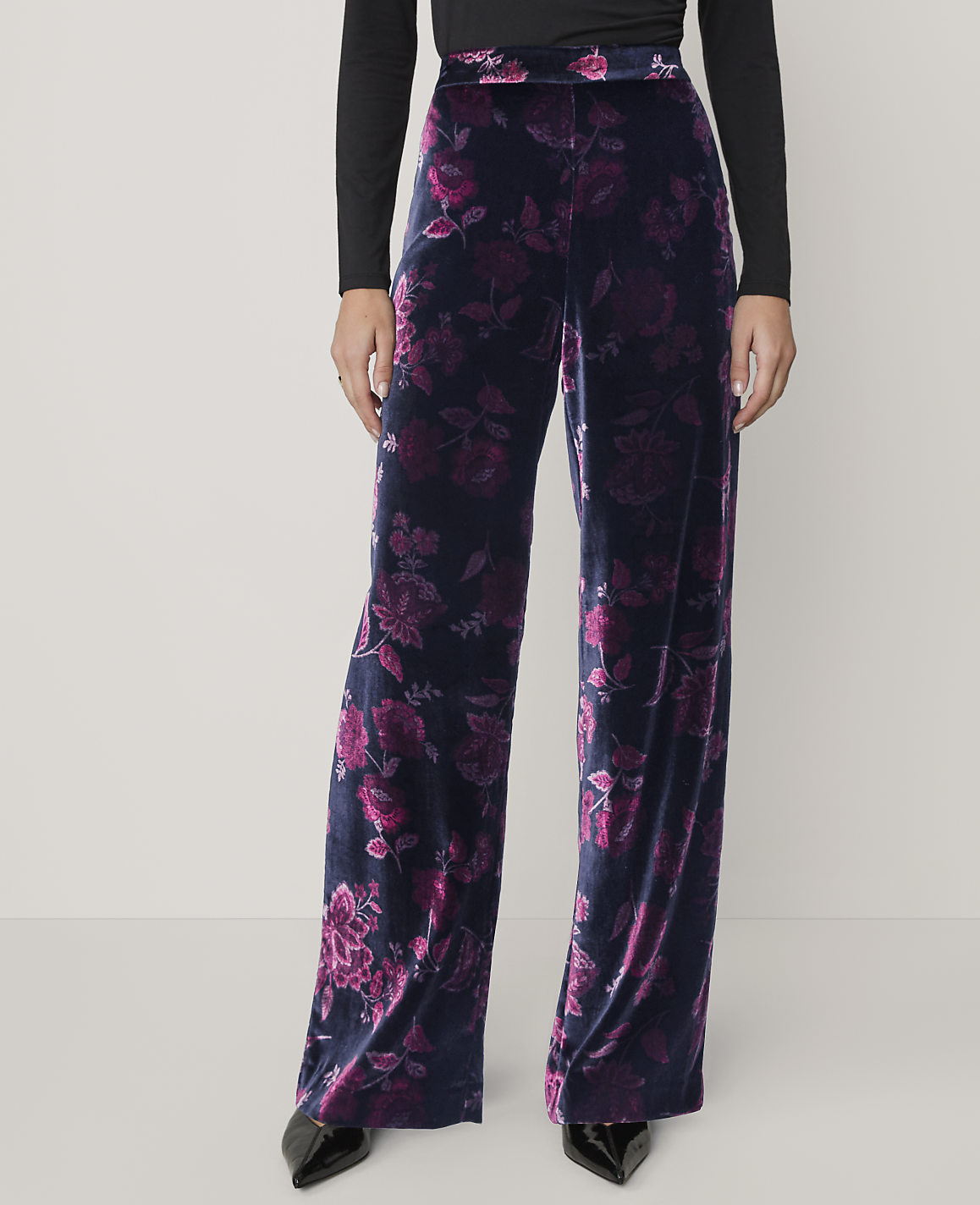 A person wears wide-leg black and purple floral velvet trousers and a black top against a plain light grey background.