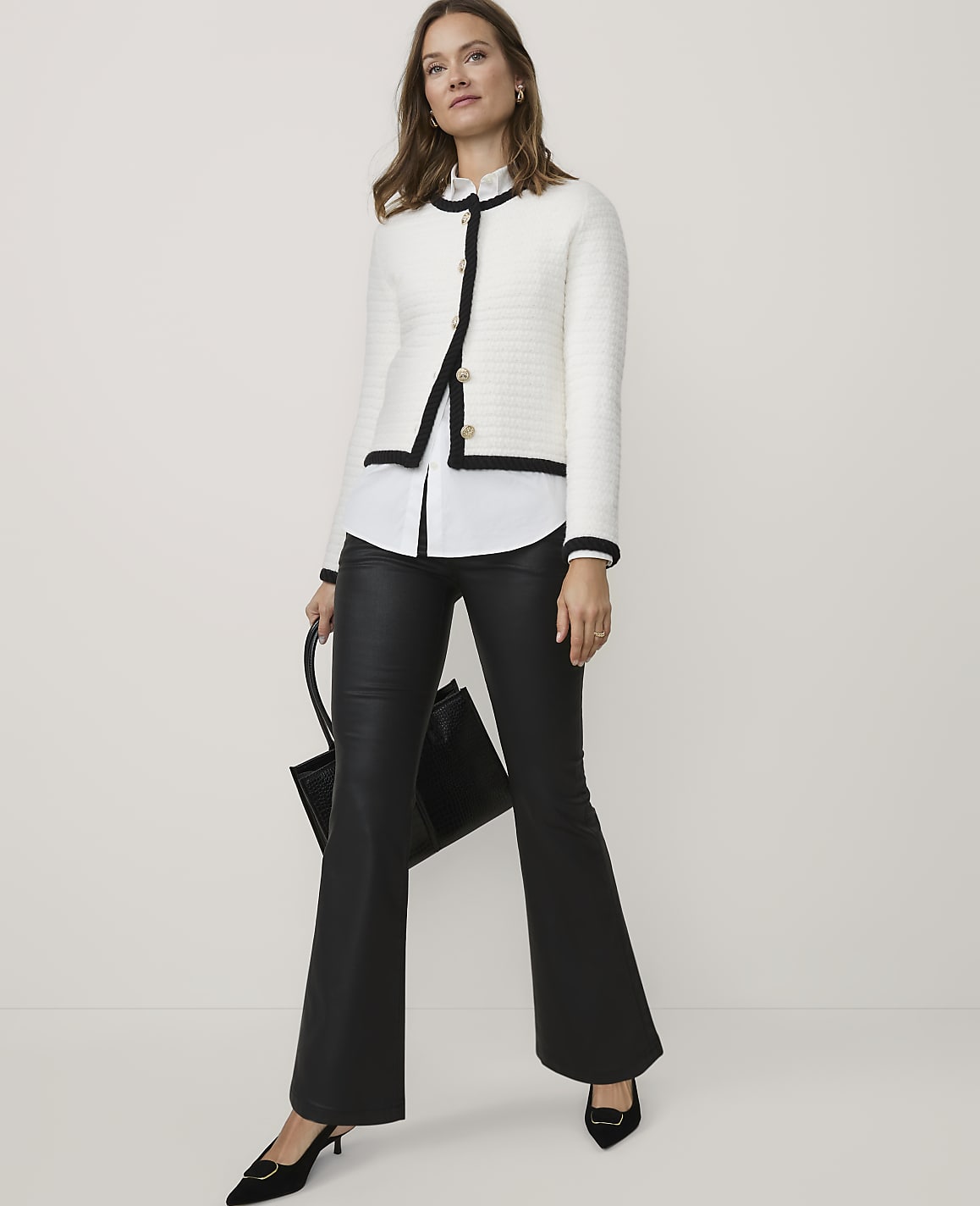 A woman wearing a white tweed jacket, white shirt, and black leather flared trousers stands against a plain white background while holding a black handbag.
