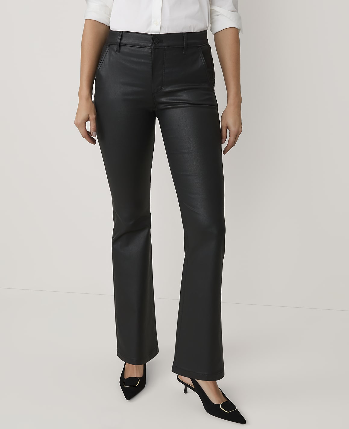 A person wearing a white shirt, black faux-leather trousers, and black pointed heels stands against a plain white background.