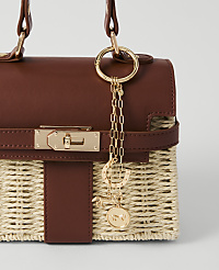A close-up of a woven straw handbag with brown leather accents and gold equestrian-themed charms hanging from the handle. carousel Product Image 1