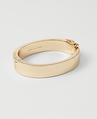 A gold bangle bracelet with a chain detail on the clasp rests on a plain white background. carousel Product Image 1