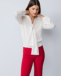 A woman in a cream bow-neck blouse and red pants stands against a plain gray background. carousel Product Image 1