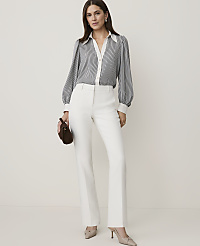A woman in a striped blouse and white pants stands against a plain, light background, holding a brown clutch. carousel Product Image 1