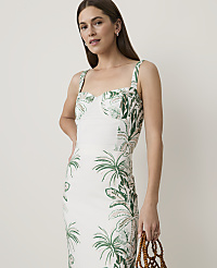 A woman in a white dress with a green tropical print stands against a plain, light background, holding a beaded bag. carousel Product Image 1