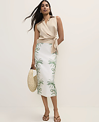 A woman in a beige wrap top and a white skirt with a palm tree print stands against a plain, light background, holding a straw bag. carousel Product Image 4