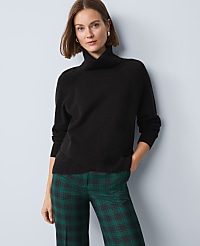 A woman in a black turtleneck sweater and green plaid pants stands against a light blue background. carousel Product Image 1