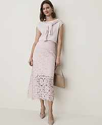 A woman in a light pink lace skirt and matching top stands against a plain, light-colored wall, holding a woven handbag. carousel Product Image 4