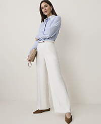 A woman in a blue striped shirt and white wide-leg pants stands against a plain, light-colored background, holding a small woven purse. carousel Product Image 1