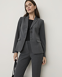 A woman in a grey suit stands against a plain, light-colored wall, holding a black bag. carousel Product Image 1