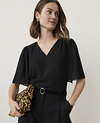 A woman in a black v-neck top and black pants holds a leopard print clutch in front of a plain background. carousel Product Image 1