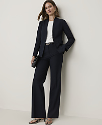 A woman in a navy pantsuit stands against a plain, light-colored wall, holding a black clutch. carousel Product Image 1