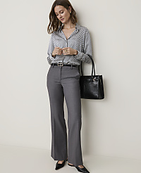 A woman in a patterned blouse and gray slacks stands against a plain wall, holding a black handbag. carousel Product Image 1