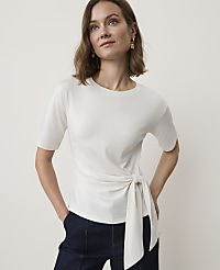 A woman in a cream-colored, tie-waist t-shirt and dark denim pants stands against a plain, light background. carousel Product Image 1