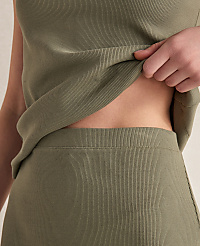 A person's midriff is visible in the foreground, with a hand lifting a ribbed olive-green top, against a plain, light-colored background. carousel Product Image 2