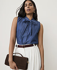 A woman in a denim bow-neck blouse and white pants holds a brown clutch in front of a plain background. carousel Product Image 1