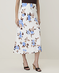 A woman models a white floral midi skirt with blue and brown flowers, paired with brown wedge sandals, against a plain, light-colored background. carousel Product Image 2