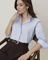 A woman in a light blue collared shirt and brown pants sits in a chair against a plain background. carousel Product Image 3