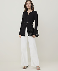 A woman in a black cardigan and white pants stands against a plain background, holding a black clutch. carousel Product Image 1