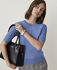 A woman in a blue sweater and plaid skirt holds a black handbag in front of a plain background. carousel Product Image 1