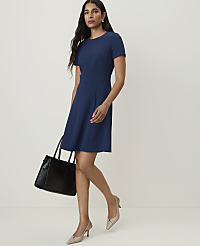 A woman in a navy blue dress and nude heels walks with a black handbag against a plain, light background. carousel Product Image 3