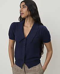A woman in a navy blue cardigan with puffed sleeves and a scalloped collar stands against a plain, light-colored background, her hands in the pockets of her plaid pants. carousel Product Image 1